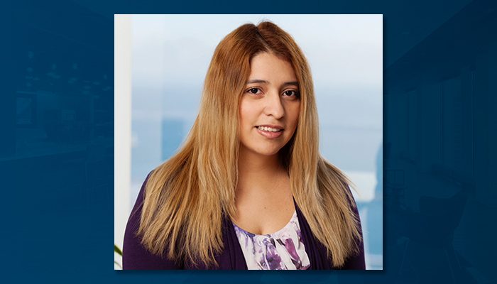 Headshot of Elizabeth Cañongo, an operations specialist with Draper and Kramer, with long, light brown hair. She is smiling softly and wearing a purple cardigan over a floral blouse, set against a bright backdrop of a blurred window with a view of the sky and water.