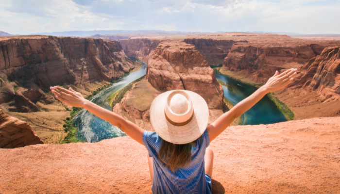 Person in a hat sitting on a rocky ledge with arms outstretched, overlooking a river winding through a canyon.