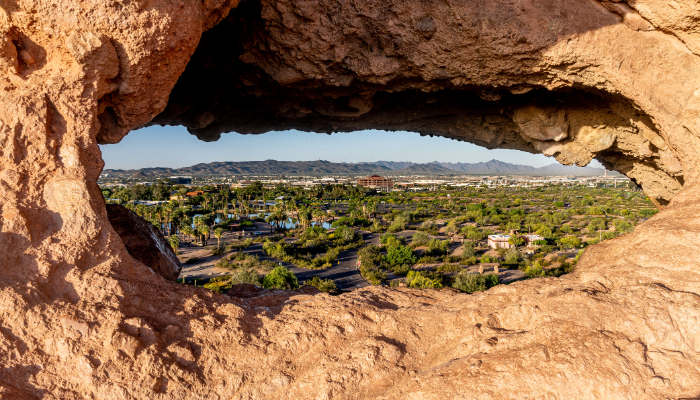 A cityscape and landscape view framed by a rocky cave opening, showing buildings, trees, and mountains in the background.