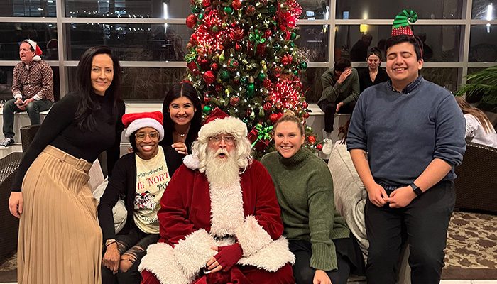 A group of people pose with Santa Claus in front of a decorated Christmas tree indoors. Some are wearing festive hats.