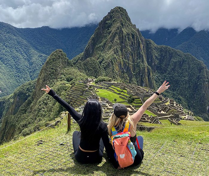 Two people with arms raised sit on grass, overlooking the ancient ruins of Machu Picchu, surrounded by lush mountains under a cloudy sky.