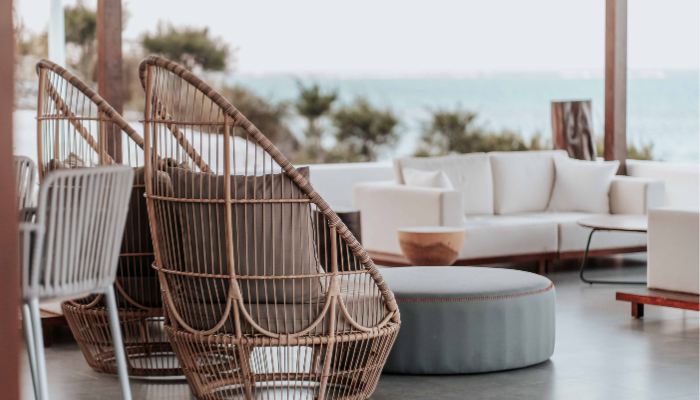 Outdoor patio with wicker chairs, grey ottoman, and white sofa overlooking the ocean with trees in the background.