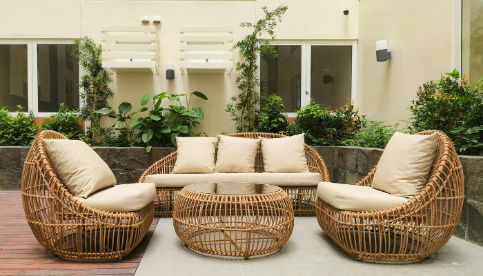 Outdoor seating area with wicker furniture, including a loveseat, two chairs with beige cushions, and a round table on a patio surrounded by greenery.