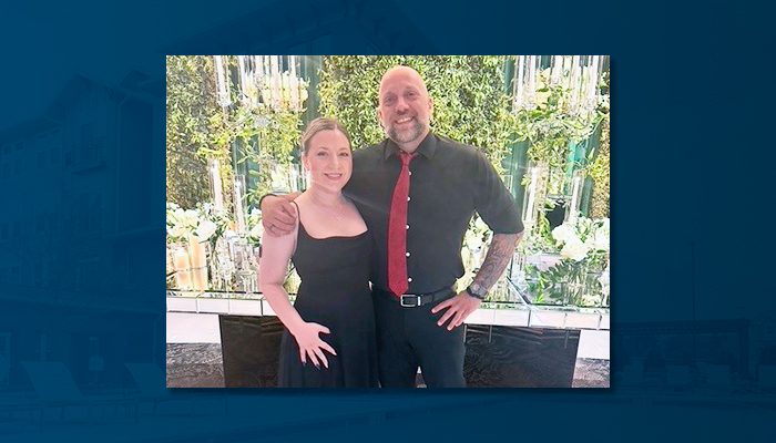 Guy and his fiancée, Eleni, standing together and smiling in front of a decorative, plant-filled backdrop. Guy is wearing a black shirt with a red tie, while Eleni wears a black dress. They have their arms around each other, looking happy.