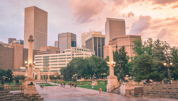 A view of downtown Denver's skyline at sunset, with modern skyscrapers surrounded by trees, statues, and historical architecture in a park-like setting.