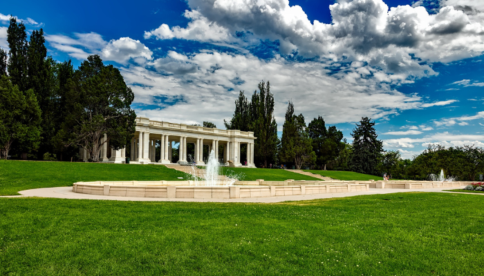 A picturesque view of a neoclassical pavilion surrounded by lush green lawns and fountains in Denver, Colorado, under a partly cloudy sky.