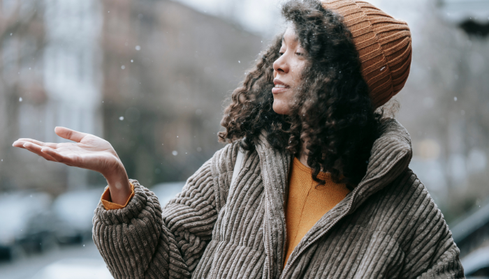 A woman wearing a warm brown beanie and a corduroy jacket enjoys the outdoors, reaching out to catch snowflakes with a smile on her face. The background features a blurred urban setting with hints of winter weather.