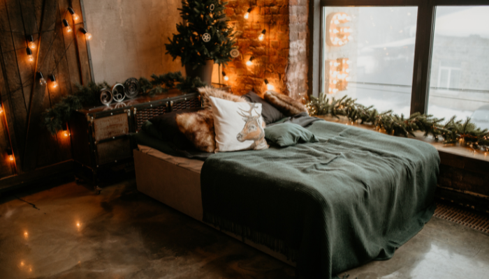 A cozy apartment bedroom decorated for winter with green blankets on a bed, a festive pillow featuring a reindeer, a small decorated Christmas tree, and warm string lights illuminating the space.