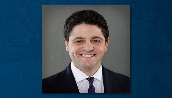 AJ Markowski, a smiling man with short dark hair, wearing a dark suit, white shirt, and purple tie, centered against a neutral gray background framed by a blue border.