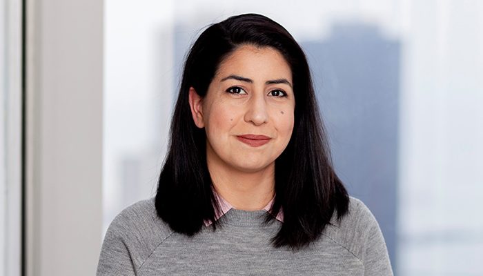 Martha Gaitan-Manning, a smiling woman with shoulder-length dark hair, wearing a gray sweater and pink-collared shirt, standing in front of a window with a blurred cityscape background.