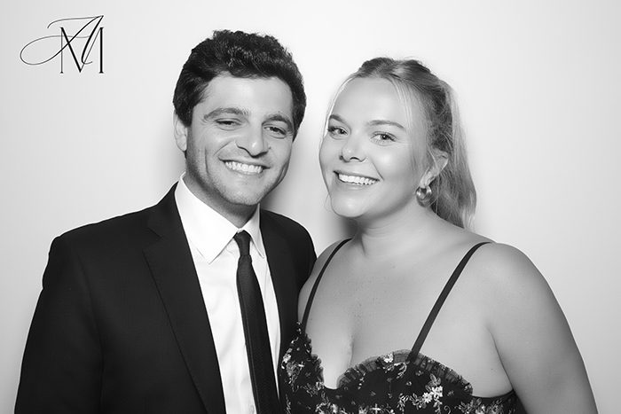 Black-and-white photo of AJ Markowski on the left, smiling in a suit and tie, standing next to his wife, Kim, on the right, who is wearing a floral dress with thin straps and smiling brightly.