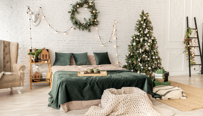 Beautifully decorated bedroom with a green bedspread, a Christmas tree, a wreath on a white brick wall, string lights, and holiday-themed accents.