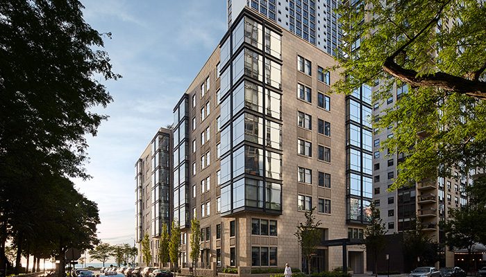 Modern apartment building in an urban neighborhood, featuring sleek glass and stone exterior design with large windows and a tree-lined street in the foreground.