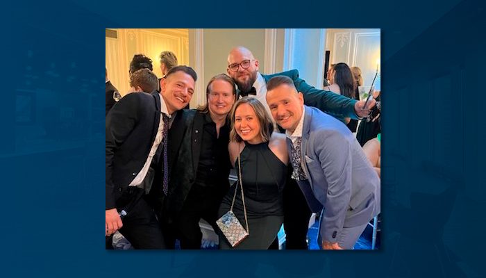 A group photo from an awards dinner featuring Heroes Salinas standing in the back, smiling with glasses and a green jacket, surrounded by coworkers and industry friends dressed in formal attire.