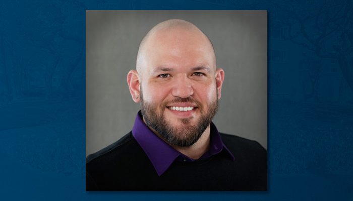 Headshot of Heroes Salinas, a smiling man with a bald head and neatly trimmed beard, wearing a black shirt with a purple collar against a neutral gray background.