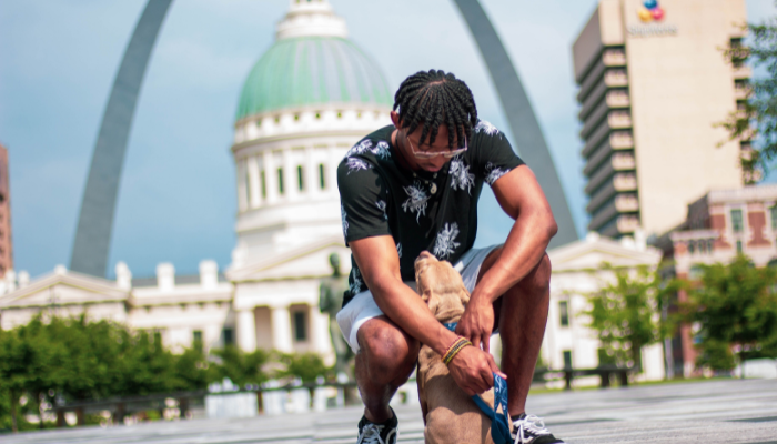 A person kneeling to pet a dog on a leash in front of the Gateway Arch in St. Louis, with the Old Courthouse visible in the background on a sunny day.