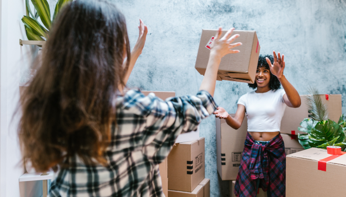 Two roommates happily tossing a cardboard box to each other while surrounded by moving boxes in a shared apartment.