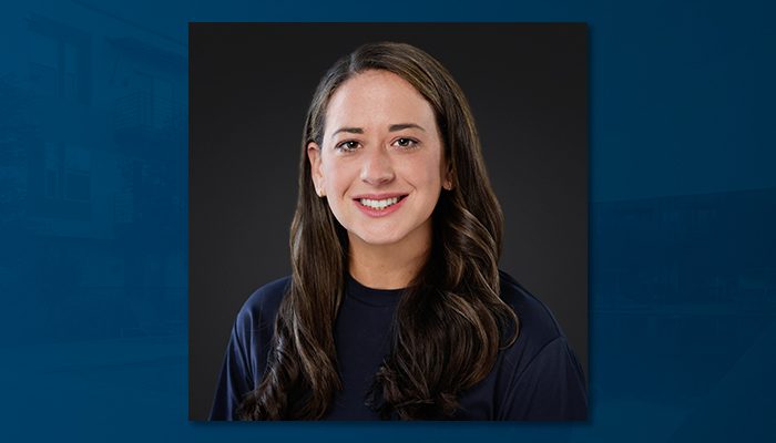 Professional portrait of Ally Shulman, a woman with long dark brown hair, wearing a navy top, smiling against a dark background.