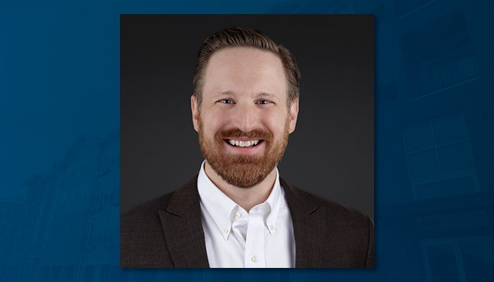 Professional portrait of Colin Volz, a man with short reddish-brown hair, a beard, and a mustache, wearing a dark blazer and white shirt, smiling against a dark background.