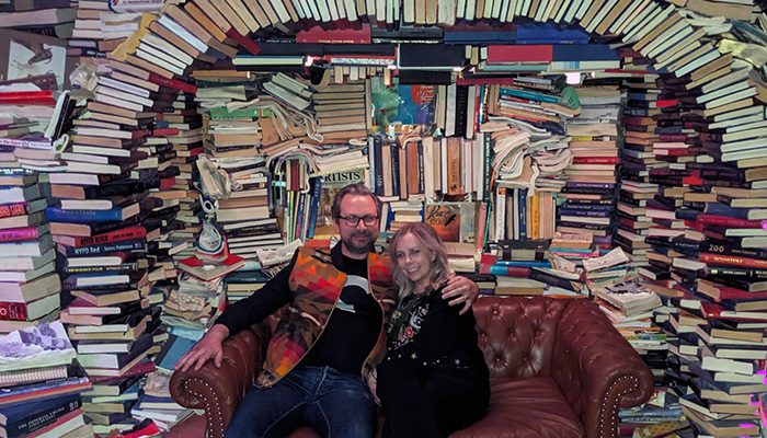 Hannah Fischer and her husband, Dorian, sitting on a brown leather couch in front of an arching wall filled with stacked books in various directions, creating a cozy and whimsical atmosphere.