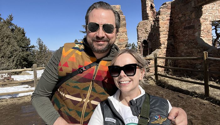 Hannah Fischer and her husband, Dorian, smiling together outdoors in front of historic stone ruins in Colorado. Hannah is wearing sunglasses and a green vest, while Dorian sports a colorful patterned vest and aviator sunglasses.