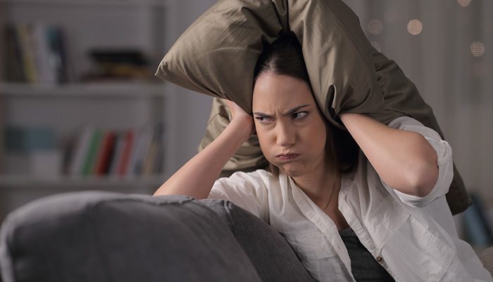 A frustrated woman sits on a couch, pressing a pillow over her ears to block out noise, illustrating the importance of being mindful of sound in shared living spaces.