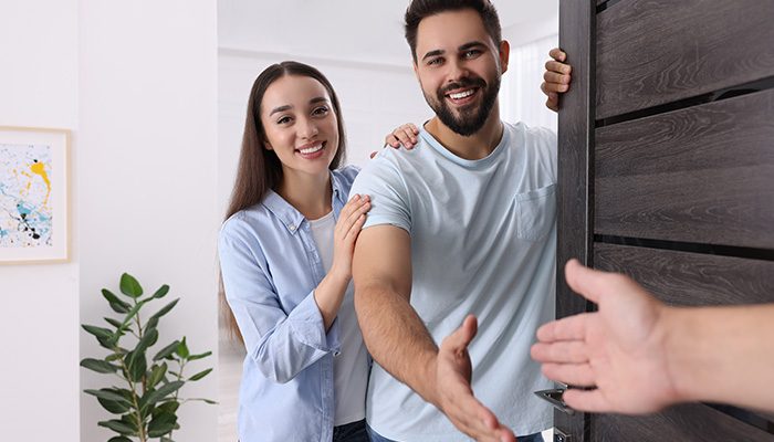 A smiling couple stands at the door of their apartment, warmly greeting a guest with an extended handshake, symbolizing neighborly kindness and a welcoming community.