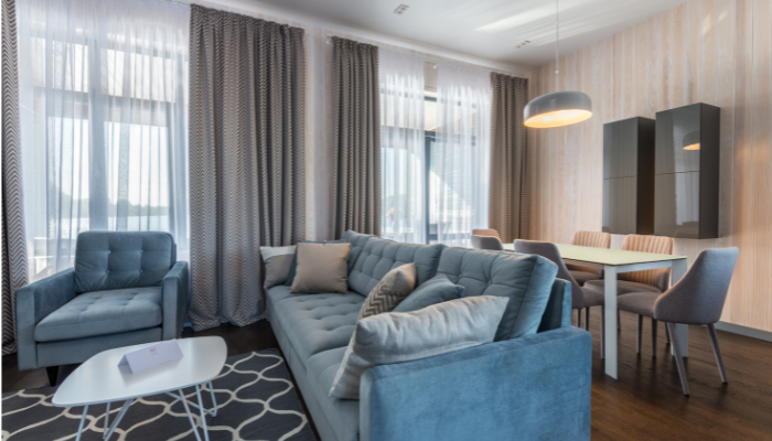 A contemporary apartment living room with a plush blue sofa, matching armchair, and a dining area with modern furnishings. The space is well-lit with natural light from large windows adorned with sheer and patterned curtains.