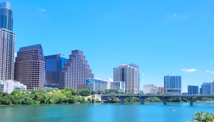 Scenic view of downtown Austin, Texas, with modern high-rise buildings, a bridge, and Lady Bird Lake in the foreground under a bright blue sky.