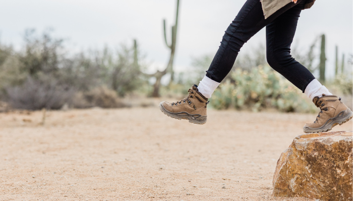 A person hiking through the desert, stepping over a rock with rugged hiking boots, surrounded by desert plants and saguaro cacti in Phoenix.