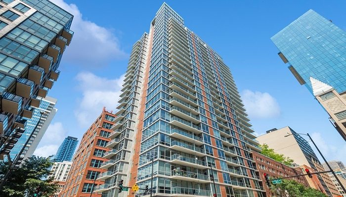 Burnham Pointe, a modern high-rise multifamily property in Chicago, featuring a sleek glass and brick facade with private balconies on each floor. The building stands tall against a bright blue sky, surrounded by other contemporary residential and office buildings in the city.