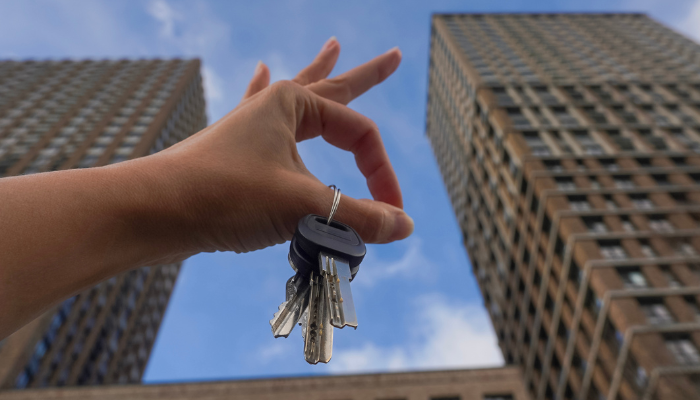 A hand holds up a set of apartment keys against the backdrop of towering residential buildings and a bright blue sky, symbolizing a successful move-in.