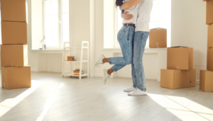 A happy couple embraces in their new apartment, surrounded by moving boxes and bathed in warm sunlight. Their joyful moment highlights the excitement of finding the perfect place to call home.