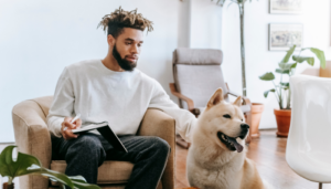 A man sits in a cozy apartment living room, writing in a notebook while petting a large, fluffy dog beside him.