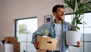 Smiling young man holding a box of household items and a potted plant while moving into a bright, modern apartment, surrounded by cardboard boxes.