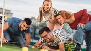 Group of young adults laughing and playing mini golf together, crouched around the putting green aiming at the hole.