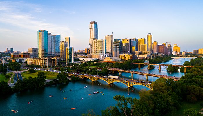 Aerial view of downtown Austin, Texas at sunset, showing the skyline, Lady Bird Lake, Congress Avenue Bridge, and people kayaking on the water.