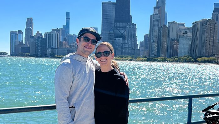 Ally Shulman and her husband, Andrew, smiling together by the lakefront with the Chicago skyline in the background on a sunny day.