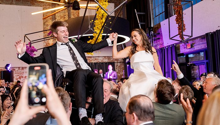 Ally Shulman and her husband, Andrew, are lifted on chairs by their wedding guests during a lively celebration, holding hands and smiling with joy.