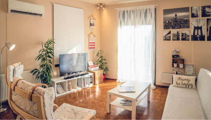 Sunny apartment living room with light-colored furniture, wall-mounted art, a compact TV stand with books and decor, indoor plants, a white coffee table with lower shelf storage, and natural light streaming through sheer curtains—illustrating tidy and functional design.