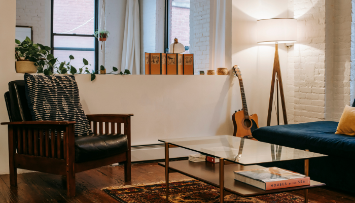 Cozy and modern apartment living room with a wooden armchair, navy blue sofa, floor lamp, indoor plants, guitar, and a glass coffee table featuring a decorative rug and books—showcasing thoughtful, space-saving design.