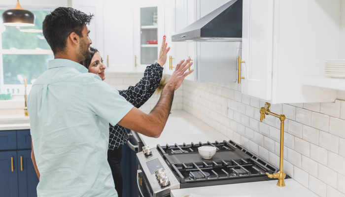 A couple tours a modern kitchen in a rental apartment, inspecting the range hood and gas stove. The kitchen features white cabinetry, brass hardware, a subway tile backsplash, and a pot filler above the stove.