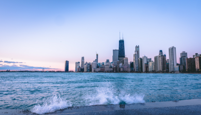 Waves from Lake Michigan splashing against the shoreline with the Chicago skyline in the background at dusk, including the John Hancock Center and other high-rise buildings.