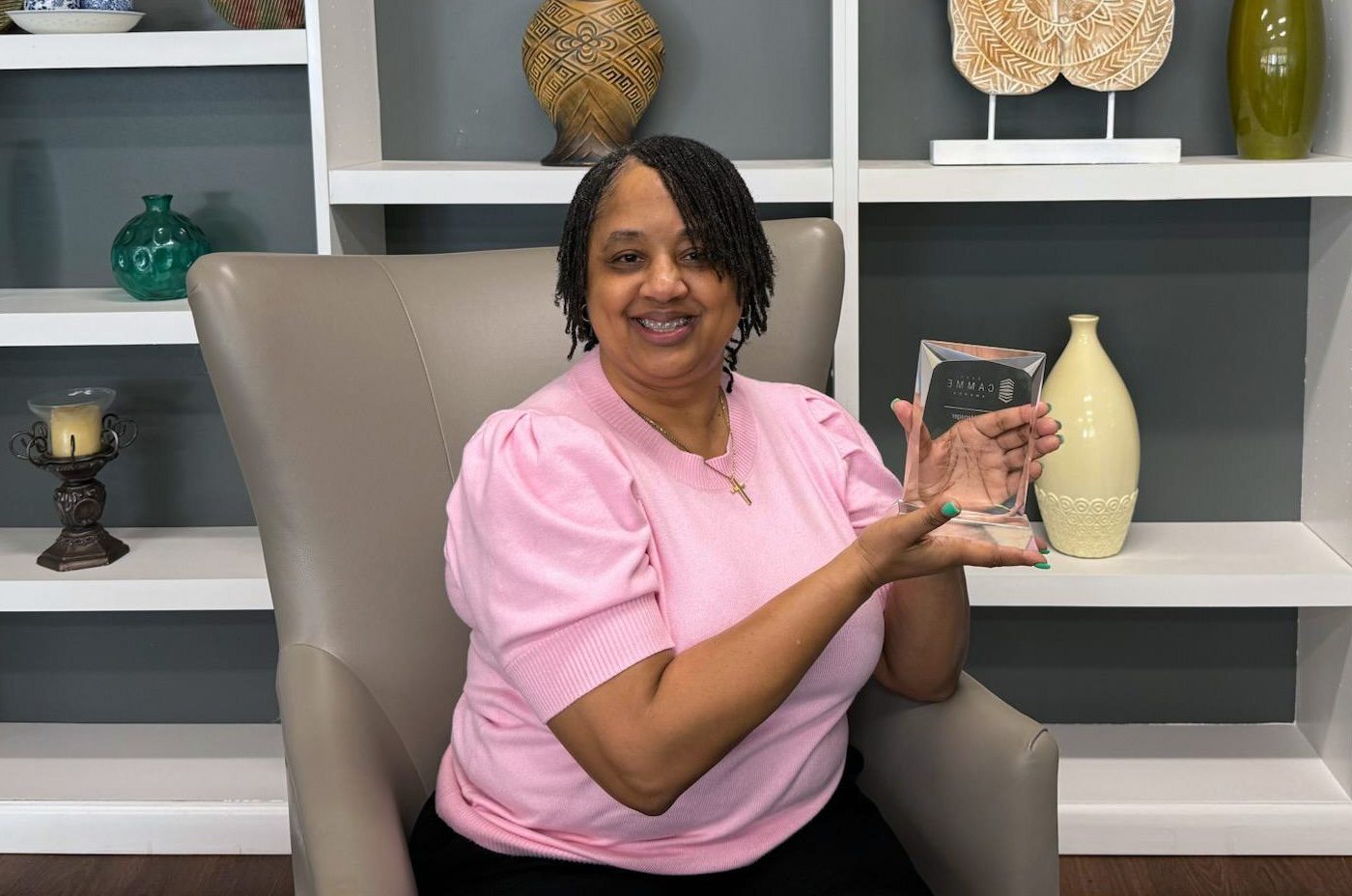 Laiche R. Gutter, seated in a modern armchair, smiles while proudly holding her 2025 CAMME Award for Property Manager in the Affordable category. She is wearing a pink blouse and sitting in front of a decorative shelving display with vases and ceramics.