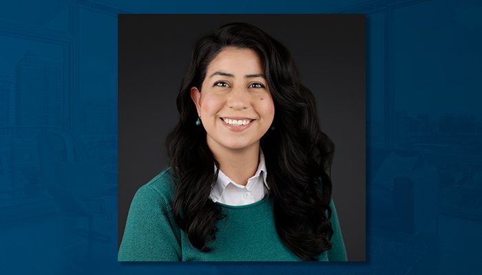 Martha Gaitan-Manning, a smiling woman with long, dark, wavy hair, wearing a green sweater over a white collared shirt, photographed against a dark background.