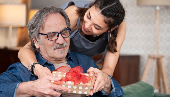 Smiling young woman giving a wrapped Father’s Day gift with a red bow to her older father, who sits on a couch in a cozy living room.