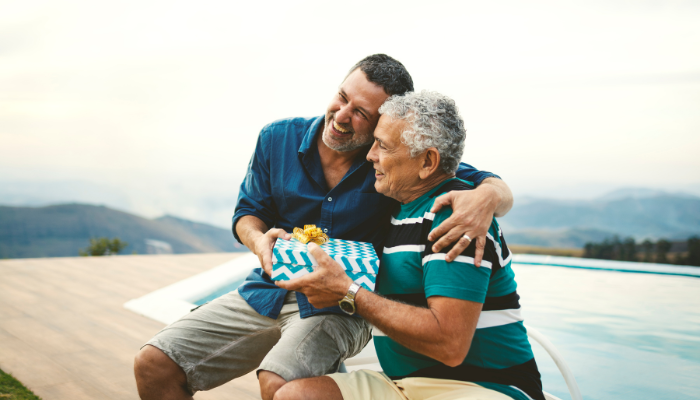 Smiling father and adult son embracing while exchanging a wrapped Father’s Day gift beside a pool with mountains in the background.