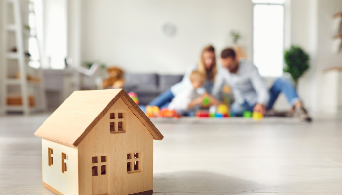 Small wooden model house on floor with blurred background of young family playing together in a bright, modern living room.