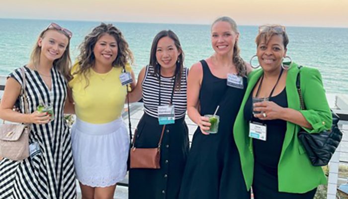 Elizabeth Soldwedel (second from right) smiling with four friends by the ocean at sunset, holding drinks and enjoying time together.