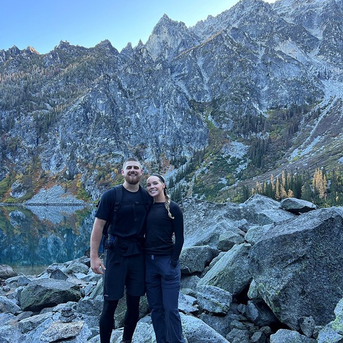 Elizabeth Soldwedel smiling with a companion while hiking in the mountains, standing on rocky terrain with a lake and tall peaks in the background.